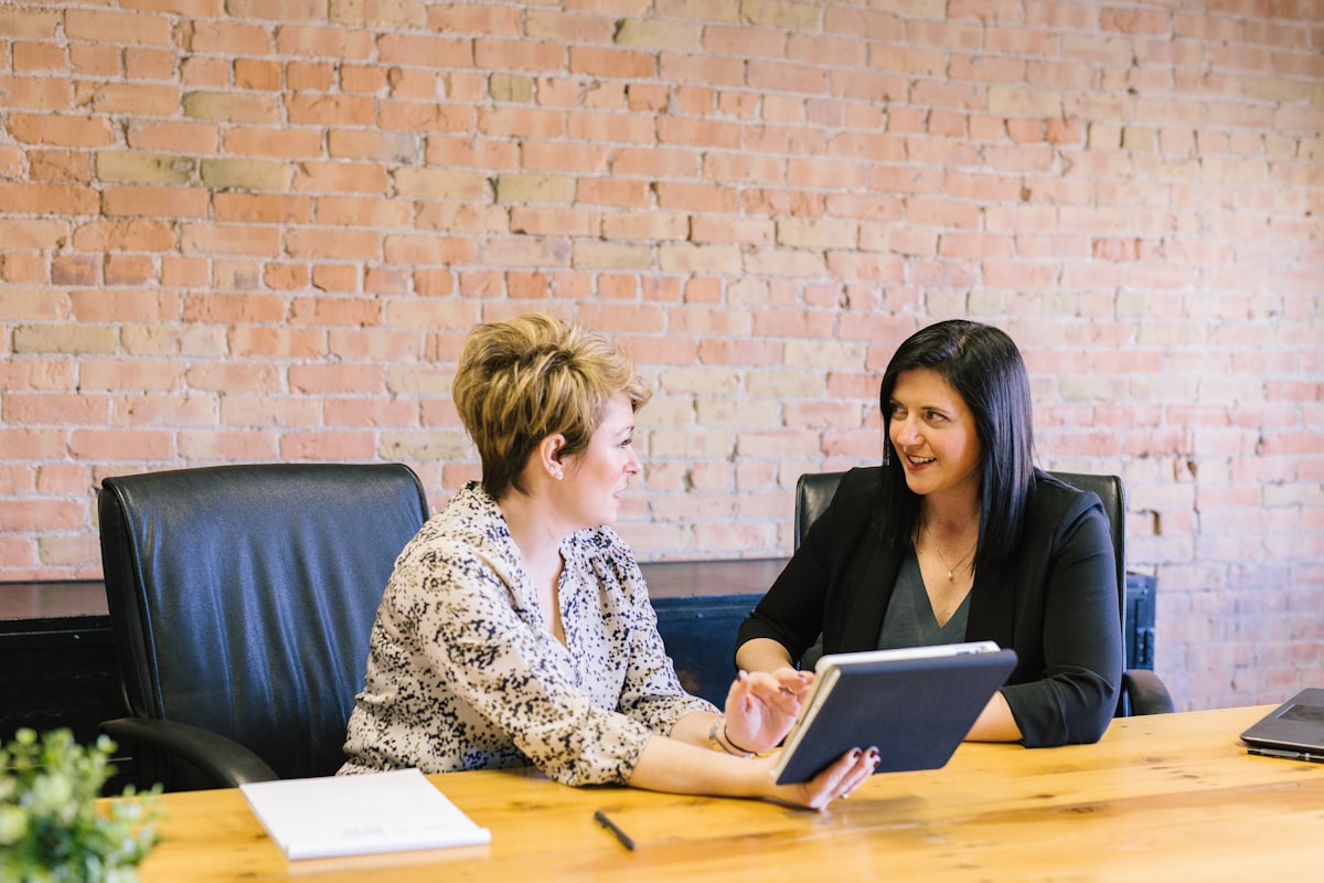 Two people in business dress talking across a meeting-room table with a laptop open between them